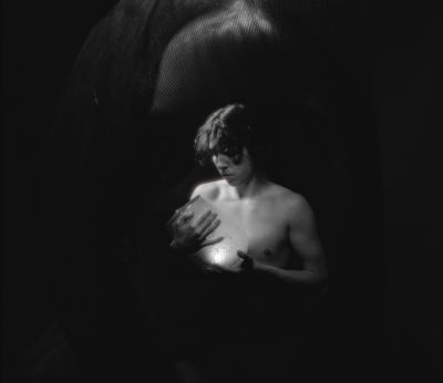 Film still from 'untitled dream film' (2024) by Jon Beaton. A young shirtless man in a black void holding a glowing vase that illuminates his chest and face.